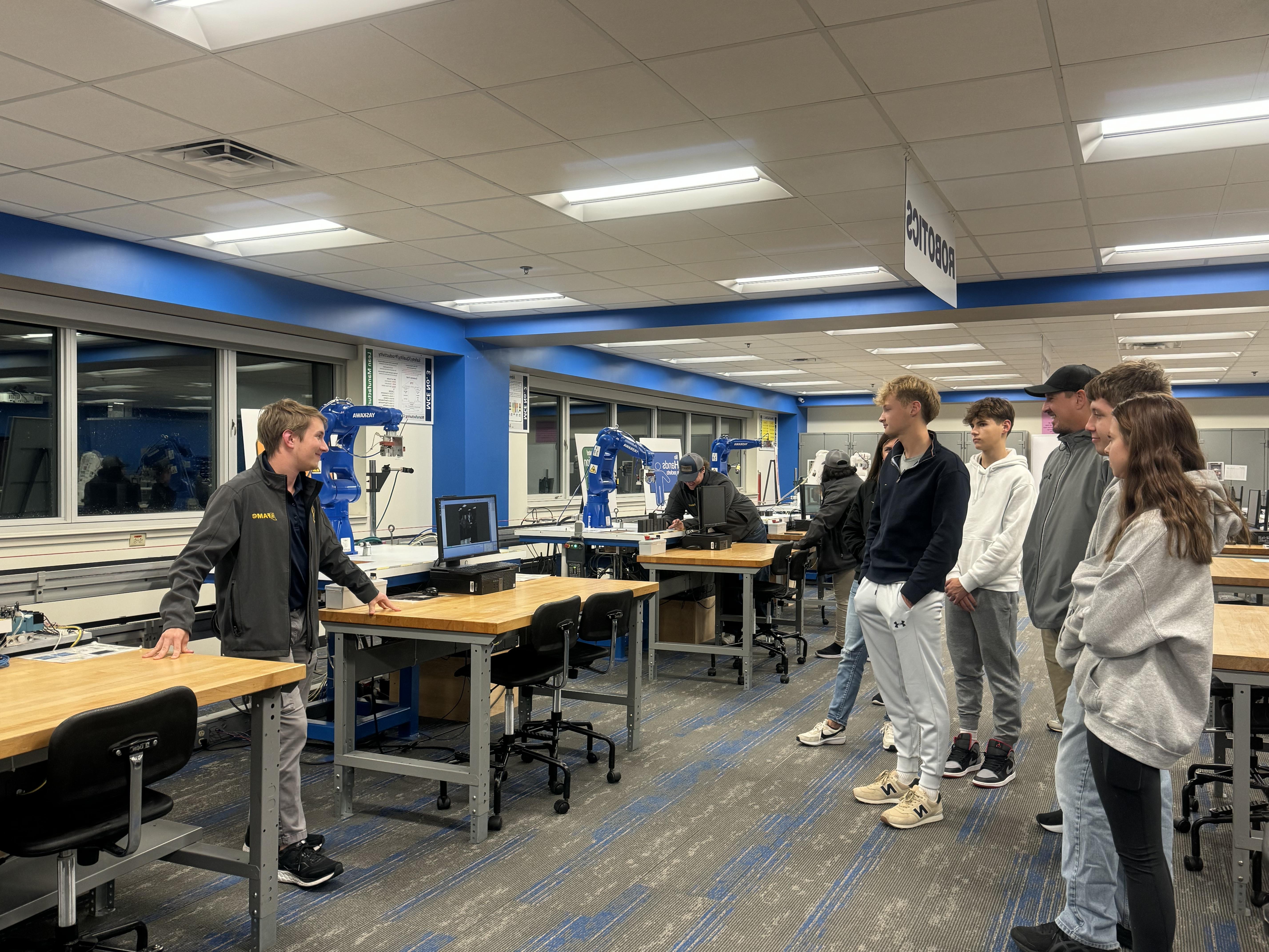 A group of male and female teens plus an adult looking at a male 澳门足球博彩官方网址 student standing next to a robotic arm. Other students and robotic arms are in the background.